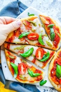 Hand picking up a slice from the margherita pizza recipe. Pizza is on a marble cutting board and white counter. 