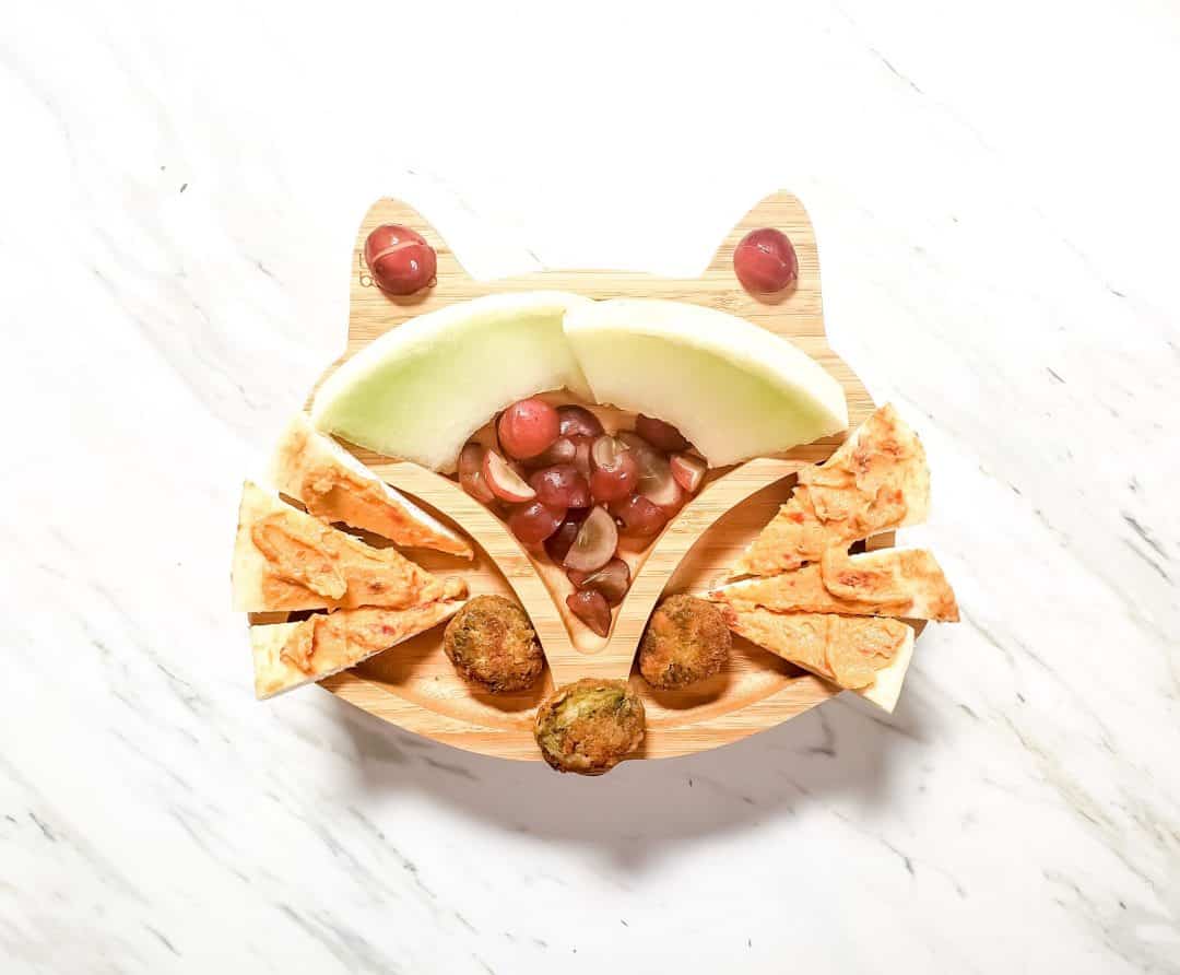 A fox plate for my son's lunch. It features honeydew, grapes, veggie nuggets and hummus on naan bread. The plate is sitting on a marble table. 