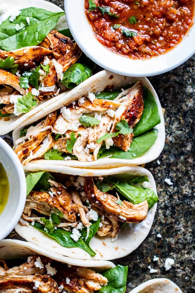 Overhead shot of grilled chicken tacos on a green stone counter with a white bowl of salsa.