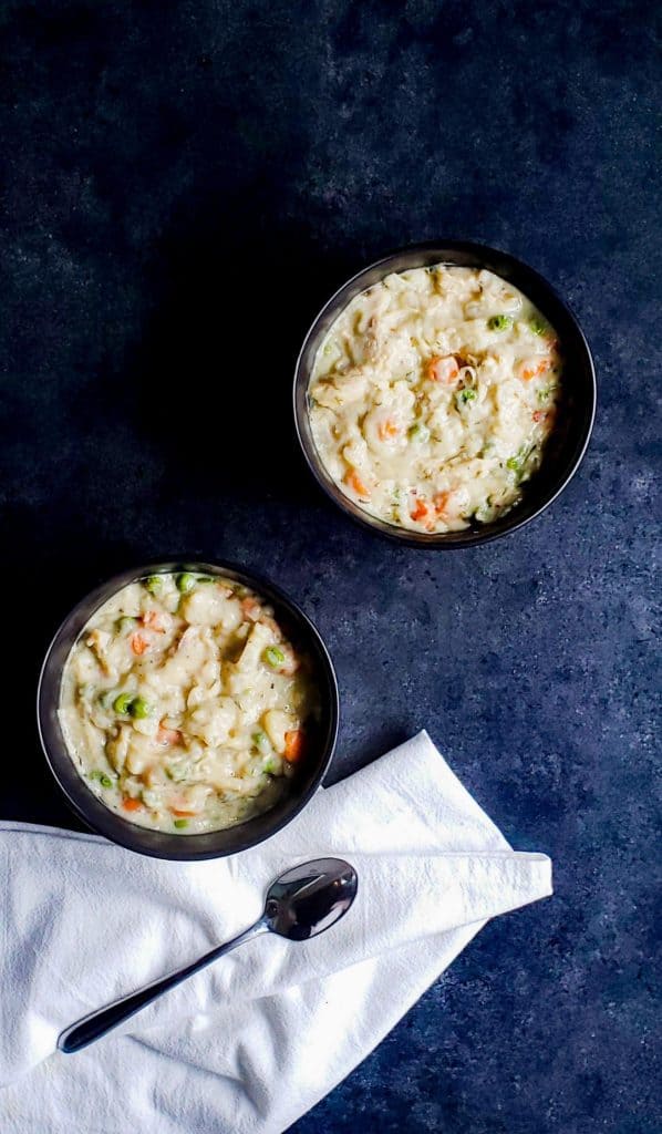 Two black bowls filled with crockpot chicken and dumplings with biscuits. The bowls are on a dark blue surface. There is a white cloth with a silver spoon on the bottom part of the photo.