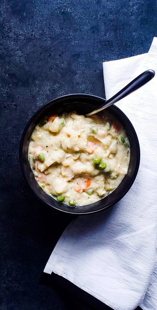 One black bowl of crockpot chicken and dumplings sitting on a dark blue counter. The shot is taken from above. There is also a silver spoon in the bowl and a white cloth on the right side.