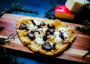 Vegetarian Flatbread Pizza on a wooden cutting board. The pizza is topped with 3 types of cheese, mushrooms, sun-dried tomatoes and fresh rosemary. The pizza is surrounded by rosemary and has cheese blocks in the background. All of this is on a blue counter top.