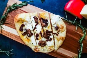 An overhead shot of the Vegetarian Flatbread Pizza recipe. The pizza is on a cutting board on a blue counter top and surrounded by fresh rosemary and 2 cheese blocks.