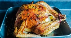 The image is a shot of herb butter roasted turkey in a silver roasting pan. The entire turkey can be seen and the shot is taken at a 45 degree angle. The pan is sitting on a blue counter top.