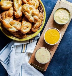 In the top left corner is a woven yellow and cream basket with homemade soft pretzel twists in it. To the left is a wooden serving tray at an angle. There are three types of cheese in the serving tray and it is oriented vertically. On bottom is a white, yellow, and blue plaid towel. All of this is shot from above on a blue counter 