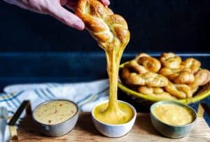 This image of homemade soft pretzel recipe is a 45 degree angle shot and has three types of cheese dips in a serving platter up front. There is a basket of pretzel twists and white towel out of focus in the background. The background and counter are dark blue. There is a hand pulling a pretzel twist out of the middle bowl and a long cheese pull hanging down. 