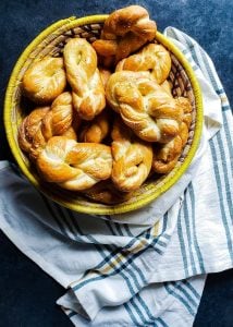 This image for the homemade pretzel recipe is of a dozen pretzel twists in a yellow and cream woven basket. The basket it in the top left corner and is sitting on a blue counter. There is a white, blue, and yellow plaid towel draped around the bottom half of the basket.