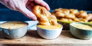 Image for pretzel sauce trio recipe. a hand on the left side dipping one of the pretzels into a white bowl of beer cheese. On the right is a green bowl of american cheese sauce. On the right is a cream bowl of chipotle queso sauce.