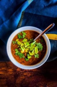 Close up above the head shot of vegan black bean soup recipe. The soup is in a white bowl with a silver spoon sticking out. The soup is topped with avocado and cilantro. The bowl is on a wooden surface with a blue, gray and yellow towel on the top half of the photo. The bowl is fully in focus in the middle of the photo.