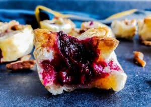 Image for blackberry brie bites with pecan sage topping recipe. The image is a close up of one bite sitting on a blue counter top. There are multiple other brie bites in the background blurred out.