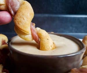 Close up strait on image for Prosciutto Wrapped Breadsticks with Gouda Cheese Dip. The image shows the twist being dipped into cheese sauce. The background in dark blue and everything is sitting on a navy counter. 