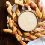 Image for Prosciutto Wrapped Breadsticks with Gouda Cheese Dip recipe. The image is an above shot. It shows a white plate with cheese dip in a gray bowl in the center of the plate. Surrounding the dip on the plate is the twists. The plate is sitting on a wooden table with a white cloth on the right side.