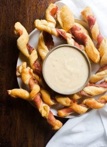 Prosciutto wrapped breadsticks on a white plate with cheese sauce in the middle on a wooden counter.