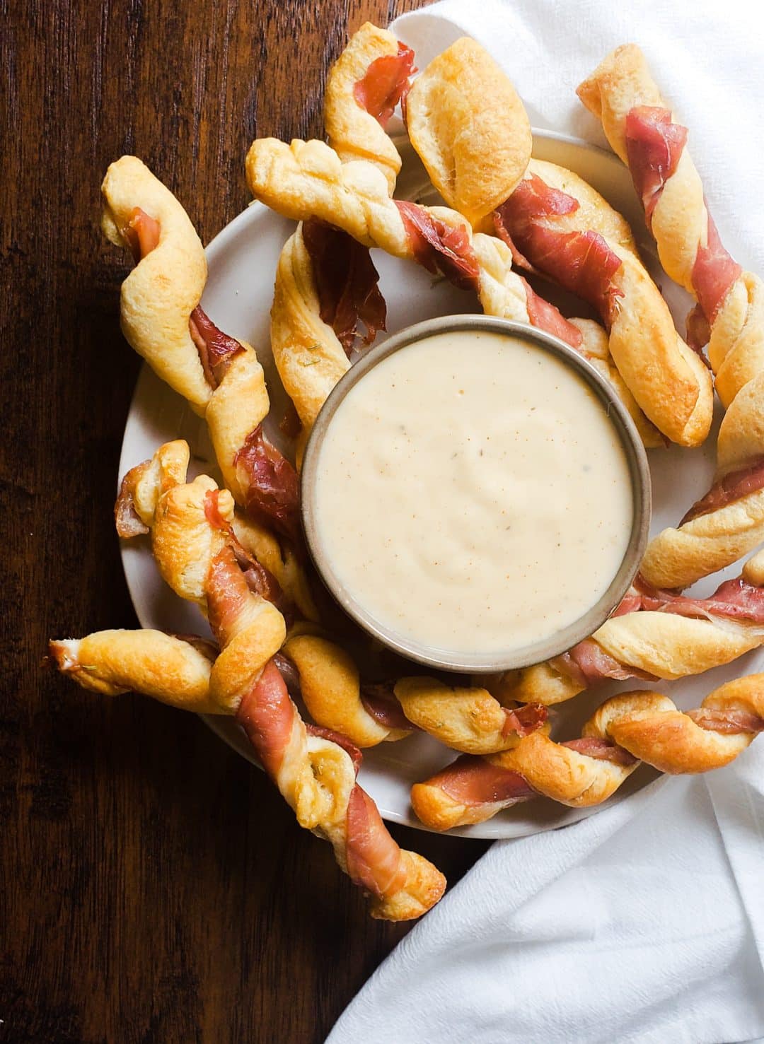 Image for Prosciutto Wrapped Breadsticks with Gouda Cheese Dip recipe. The image is an above shot. It shows a white plate with cheese dip in a gray bowl in the center of the plate. Surrounding the dip on the plate is the twists. The plate is sitting on a wooden table with a white cloth on the right side.