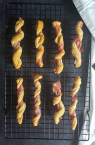 Prep image for Prosciutto Wrapped Breadsticks with Gouda Cheese Dip. The image shows the finished bread twists cooling on a black backing wire rack. The rack is sitting on a dark brown counter top and there is a white cloth on the right side.