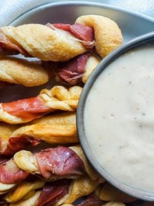 Extreme close up image for Prosciutto Wrapped Breadsticks with Gouda Cheese Dip. The image is an extreme close up. On the right side half out of frame is cheese dip in a gray bowl. On the left side of the dip is bread twists on a white plate. There is a white cloth above the plate out of frame. The image is shot from above.