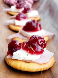 Image for Ritz Bites with Ham, Cranberries and Brie. Image is strait on and shows the bites in a line on a wooden surface. The front one is in focus with the rest in the back out of focus.