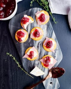 Image for Cranberry Brie and Ham Ritz Bites. Image is shot from above and shows bites on a blue marble serving tray on a black surface. There is also thyme leaves spread out with a white bowl of cranberry sauce on the top left. 
