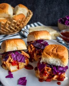 Image for Crockpot Hawaiian BBQ Chicken Sliders. Image shows 2 sliders on a white plate. In the background is a basket of rolls, cheese slices, sauce and cabbage. 