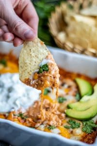 Shot of a chip being dipped into the chicken enchilada dip by a hand. There is a basket of chips blurred in the background. 