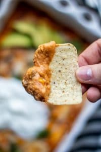 Close up shot of the chicken enchilada dip on a tortilla dip being held. The entire baking dish is blurred in the background. 