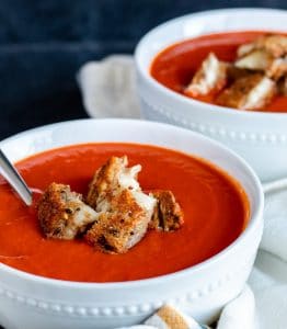 Roasted red pepper tomato soup in a white bowl with silver spoon in it. Plus grilled cheese bites on top.