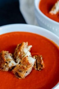 Close up shot of soup. The shot is close up and focuses on the front white bowl of soup. There are grilled cheese bites on top. There is also a second bowl faded in the background.