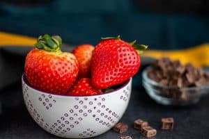 A white bowl of strawberries with a bowl of chocolate chips in the background blurred out. These both are sitting on a dark blue counter. In the background is blue and yellow.