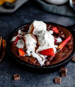 A close up shot of the skillet brownie in an iron skillet topped with vanilla ice cream, hot fudge, and strawberries. There is a tiny peek of gold silverware on the left out of frame. This is all sitting on a dark blue counter.