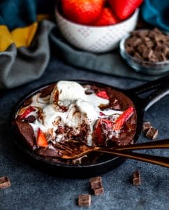 45 degree angle shot of skillet brownie . The skillet is sitting on a blue counter and have a gold fork and spoon it it with some of it eaten. In the background is a bowl of strawberries, chocolate chips, a clue and grey towel blurred out. 