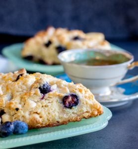 Strait on shot of scone on a teal plate with second blurred in the background with a white tea cup. 