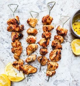 Overhead shot of chicken on four skewers on a white marble counter with lemons on the side. 