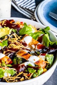 Shot of salad in white bowl with plate and dressing out of frame in the back.