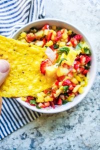 Tortilla chip being held with salsa on it and white bowl blurred underneath. 