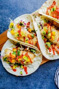 Overhead shot of healthy fish tacos on a wooden cutting board on a blue counter. 