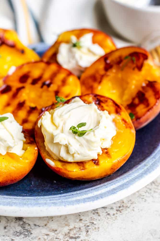 Close up of grilled peaches on a blue plate with mascarpone cheese and thyme on top. 