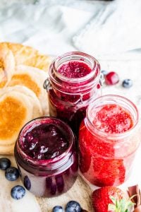 Strawberry, blueberry, and cherry jam in glass containers with fruit around them.