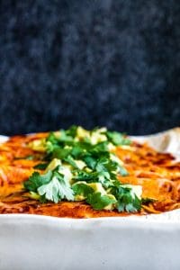 Close up shot of enchiladas in a white dish with avocado and cilantro on top.