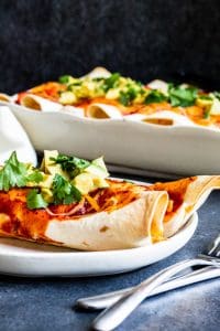 Shot of two veggie enchiladas on a white plate with silver fork and knife on the side. A white baking dish with more in the background.