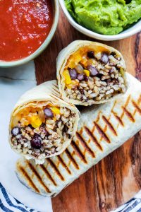 Overhead shot of burritos on a wooden cutting board with bowls of salsa and guacamole next to it.