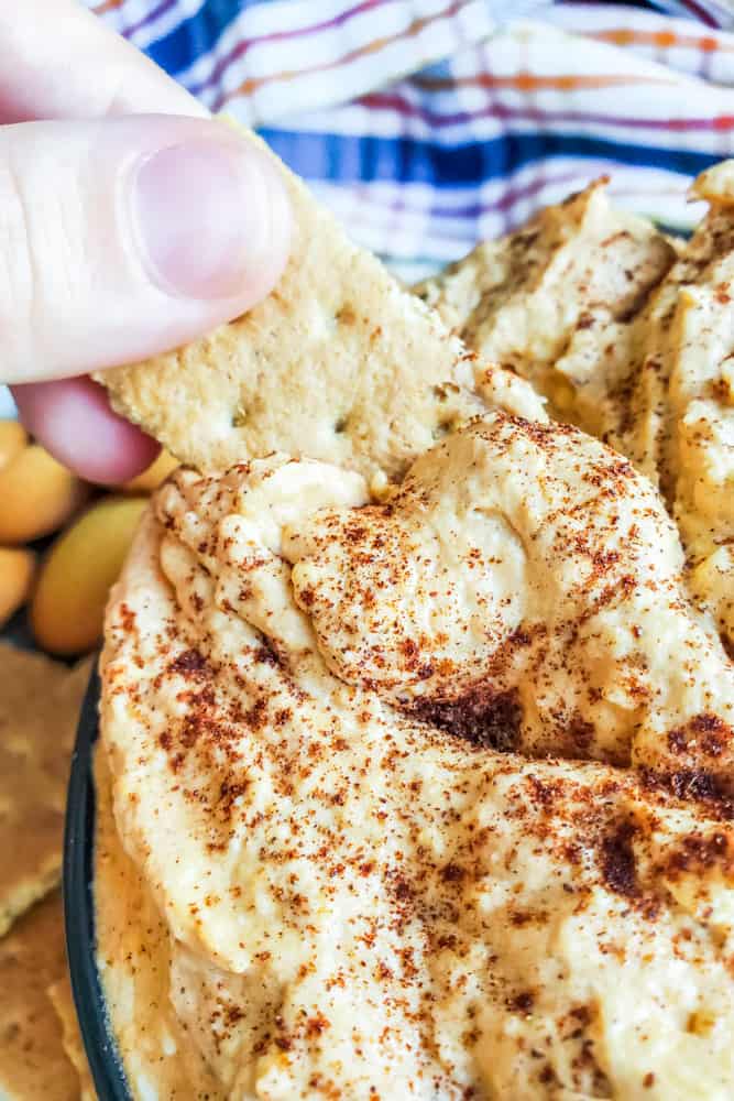 Hand dipping cracker into dip with plaid towel in the background. 