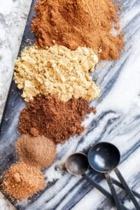 Cinnamon, nutmeg, ginger, cloves and allspice on a blue marble board in a line with black measuring spoons next to them