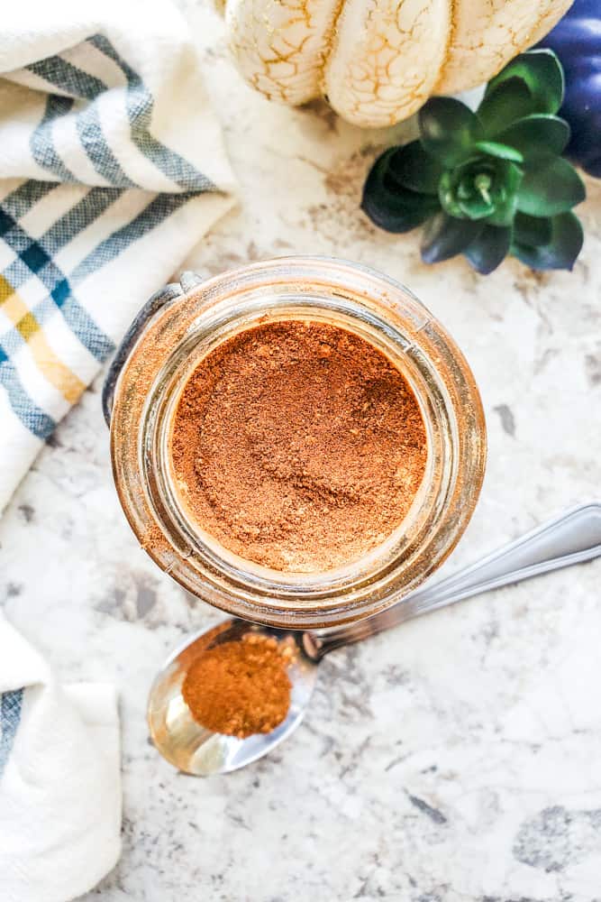 Overhead shot of pumpkin spice in a glass jar on a white counter with silver spoon next to it.