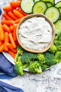 Overhead shot of ranch dip surrounded by veggies on a white plate.
