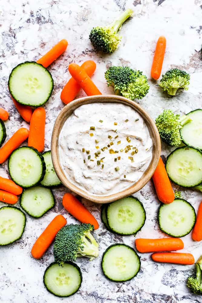 Ranch in a gray bowl with veggies surrounding it on a white counter.