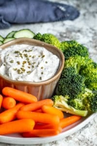 Ranch dip in a gray bowl surrounded by veggies on a white counter with blue towel in background.