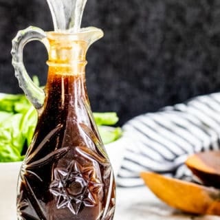 Glass bottle of balsamic vinaigrette on a white counter with salad in the background.