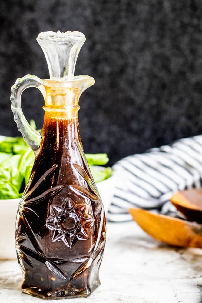 Glass bottle of balsamic vinaigrette on a white counter with salad in the background.