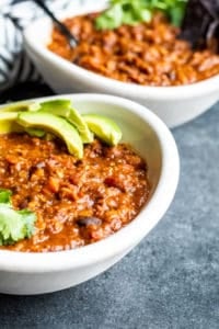 Two white bowls of vegetarian chili on a blue counter with cilantro and avocado on top.