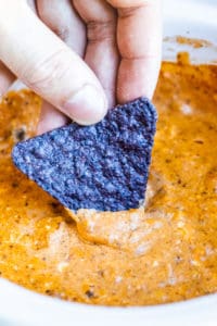 Hand dipping blue tortilla chip into chili cheese dip.